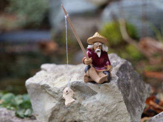 Keramikfigur sitzender Fischer mit Angel und Fisch, rot gekleidet, auf Stein am Teich – dekorative Miniteich-Figur für Garten und Balkon